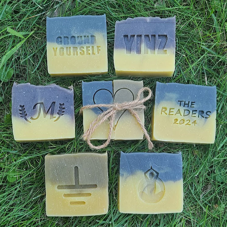 Six handmade rectangular soap bars arranged on green grass, each with a different debossed design or text including 'GROUND YOURSELF', 'VINZ', 'M', 'THE READERS 2024', a grounding symbol, and a teardrop symbol. Two soaps are blue-gray and yellow, and one soap is tied with a twine bow.
